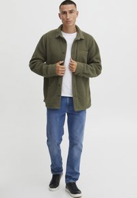Grønn fleecejakke med krage, knappestengning og brystlomme, båret over en hvit t-skjorte og blå jeans med svarte joggesko.