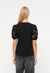 Μαύρο μπλουζάκι με διάφανα φουσκωτά μανίκια κεντημένα με λουλούδια, στρογγυλή λαιμόκοψη και μονόχρωμη πλάτη. Συνδυάζεται με σκούρα γκρι τζιν με λογότυπο σε πατς.