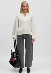 Weißer Strickpullover mit tiefem V-Ausschnitt und gerippter Textur, kombiniert mit hochgeschnittenen grauen weiten Jeans und einer großen glänzenden schwarzen Tragetasche.