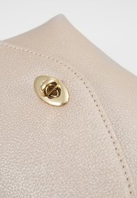 Angolo di una borsa in pelle beige con texture, caratterizzato da un fermaglio ovale dorato e una sottile cucitura decorativa.