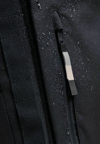 Černý voděodolný batoh z látky s texturovaným povrchem, vybavený zipovým uzávěrem a kapkami vody na materiálu. Důraz na detail stahovacího zipu.