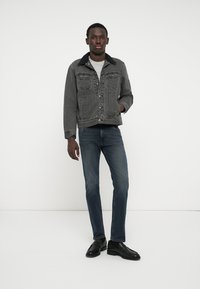 Bărbat în picioare, îmbrăcat cu o jachetă din denim neagră cu guler de blănuri, cămașă gri, blugi bleumarin și mocasini din piele negri, pe un fundal alb.