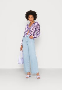 Blusa roxa e branca com estampado e laços, jeans de perna larga azul claro, bolsa lavanda e sandálias de salto roxo claro.