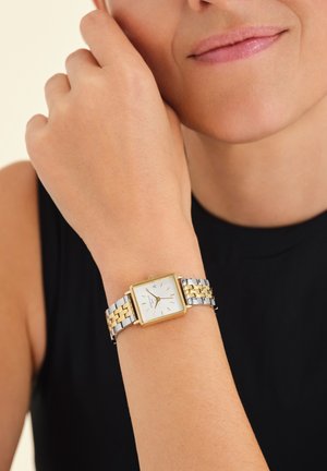 Montre bracelet carrée avec un cadran blanc, des accents dorés, et un bracelet en acier inoxydable comprenant des maillons dorés et argentés. Surface lisse et polie.
