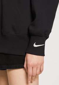 Černá mikina z bavlněné směsi s žebrovanými lemy. Bílé logo Nike Swoosh na levém zápěstí. Hladká textura, volný střih a minimalistický design.