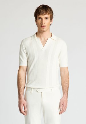 Giovane uomo con capelli castani corti che indossa una maglia bianca a maniche corte in maglia e pantaloni bianchi abbinati, in piedi davanti a uno sfondo semplice.