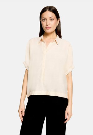 Femme aux cheveux raides et foncés porte un chemisier ample beige clair à manches courtes et un pantalon noir, debout devant un fond gris clair uni.