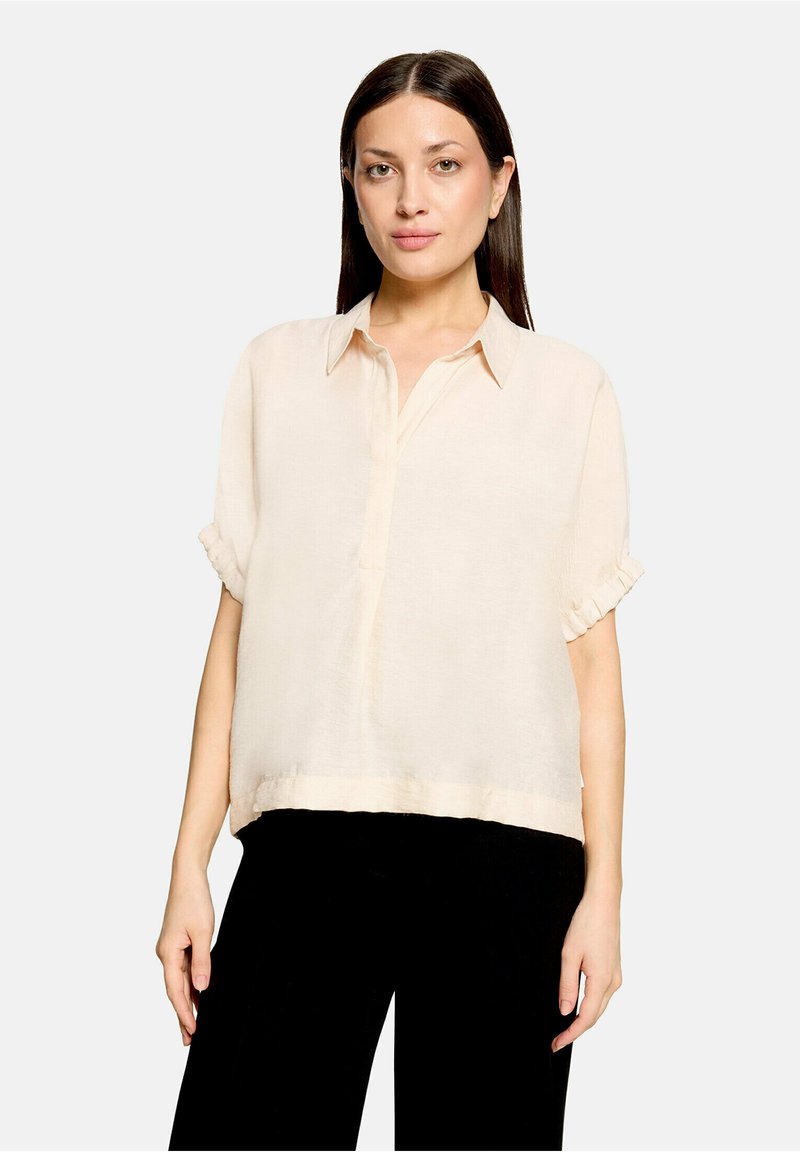 Femme aux cheveux raides et foncés porte un chemisier ample beige clair à manches courtes et un pantalon noir, debout devant un fond gris clair uni.