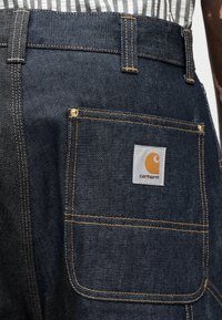 Κοντινή λήψη πίσω τσέπης από σκούρο μπλε τζιν με πορτοκαλί ραφές και λευκό λογότυπο Carhartt στο έμπλαστρο της τσέπης.