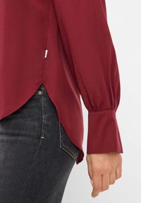 Maroon long-sleeve shirt with a slightly curved hem, gathered cuffs, and a small label at the side, paired with dark jeans.