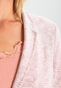 Lichtroze vest met een gestructureerd patroon en een open voorkant. Kenmerkt zich door zacht materiaal en een ontspannen pasvorm, gedragen over een bijpassend topje.