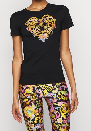 T-shirt noir ajusté avec un cœur orné de dorures et le texte « Versace Jeans Couture », associé à des leggings colorés au motif baroque.