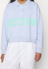 Ljusblå kortärmad sweatshirt med en mintgrön horisontell rand och "adidas"-logotyp. Den har en polo-krage och ribbad nederkant.