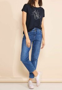 Námořnické tričko s krátkým rukávem a bílou grafikou, spojené se světle modrými džíny rovného střihu a teniskami s růžovými akcenty, na béžovém pozadí.