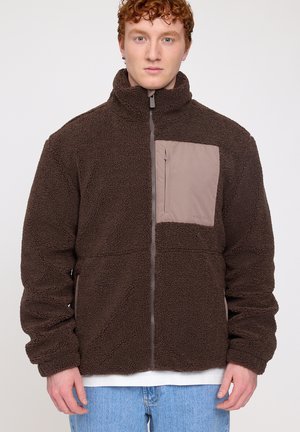Braune Fleecejacke mit hohem Kragen, Reißverschluss vorne und beiger Brusttasche. Der Stoff hat eine weiche, strukturierte Optik, die sich gut für Freizeitkleidung eignet.