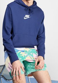Marineblå hoodie med framficka och dragsko i huven, med en vit Nike-logotyp; ihopparad med färgglada mönstrade shorts i olika pastellfärger.