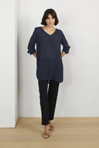Tunika lunga blu navy con scollo a V, maniche a tre quarti, tasca frontale, abbinata a pantaloni neri e sandali beige con cinturini su un pavimento di legno.