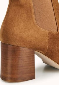 Bottine en daim marron avec des panneaux élastiques latéraux et un talon épais en bois, présentant une texture lisse et des lignes épurées.