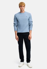 Jasnoniebieski sweter z dzianiny w warkocze z prążkowanymi mankietami, noszony z granatowymi spodniami i białymi sneakersami. Posiada subtelną naszywkę na piersi.