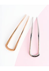 Kaksi metallista hiusneulaa vierekkäin, toinen kultasävyinen ja toinen hopeasävyinen, lepäämässä valkoisella ja vaaleanpunaisella taustalla.