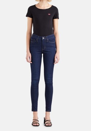 Kobieta w czarnej bluzce z krótkim rękawem z czerwonym logo, ciemnoniebieskich obcisłych dżinsach z wysokim stanem i czarnych sandałach na obcasie z paskami, stojąca.