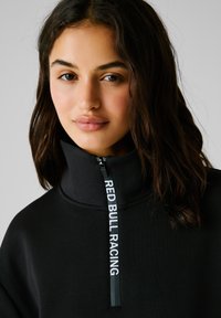 Sudadera con cuello alto y cremallera negra con "RED BULL RACING" impreso en la cremallera. Tejido suave, diseño casual y corte holgado.