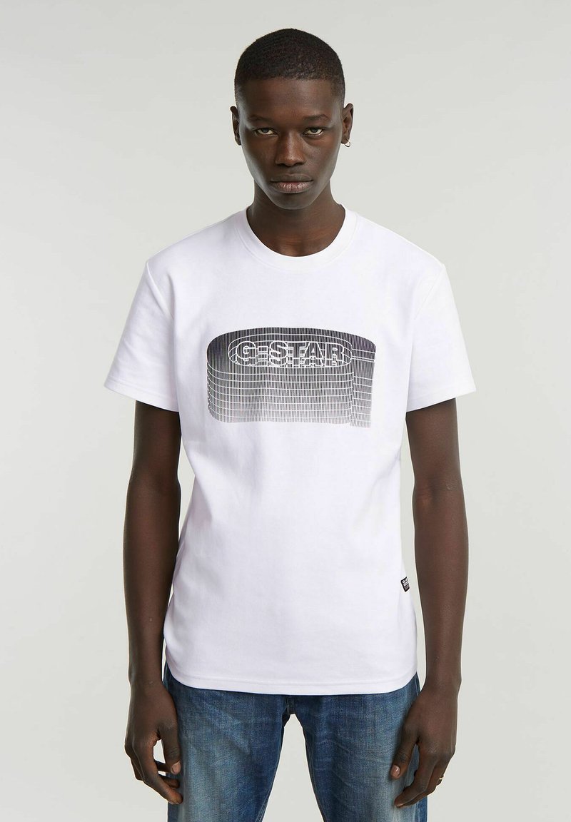 Joven con una camiseta blanca con un logo "G-STAR" negro en estilo 3D, combinado con jeans azules, de pie contra un fondo liso.