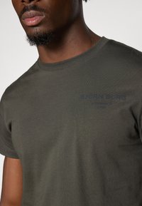 Donkergroene katoenen T-shirt met korte mouwen, voorzien van een ronde halslijn en subtiele blauwe grafische tekst op de borst die "BJÖRN BORG" leest.