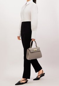 Borsa grigia con un design strutturato, superficie texturizzata e dettagli in oro. Il modello indossa una blusa bianca e pantaloni neri, mostrando un look elegante.