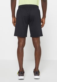 Czarne sportowe spodenki z elastycznym pasem, o gładkiej fakturze i długości do połowy uda. Noszone z szaro-żółtymi butami sportowymi.