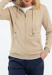 Hoodie zippé beige avec un tricot texturé, cordons de serrage, poches latérales et poignets côtelés. Dispose d'un petit logo sur la poitrine.