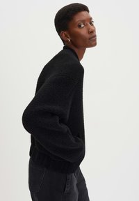 Maglione nero a maglia con superficie testurizzata, orlo a coste e maniche oversize, indossato con pantaloni scuri su uno sfondo chiaro.