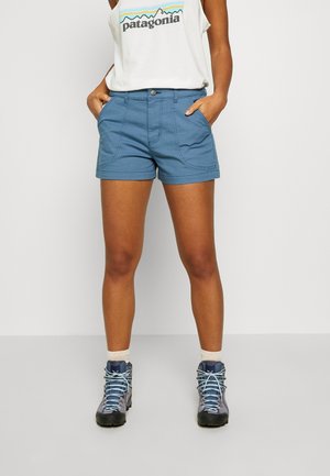 Persoon die een witte Patagonia tanktop, blauwe korte broek met zakken, witte sokken en grijze wandelschoenen draagt, staand tegen een effen achtergrond.