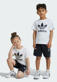 Beli bombažni majici s črnim logotipom Adidas, črne kratke hlače z belimi stranskimi črtami in bele teniske s črnimi detajli.