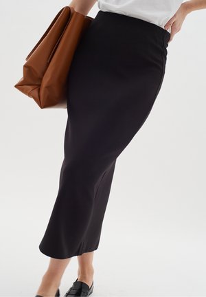 Femme portant une jupe crayon midi noire, un top blanc, des mocassins noirs, tenant un grand sac à main en cuir marron sur un fond uni.
