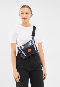 Svart axelremsväska med röd logotyp, justerbar rem, genomskinlig framficka och blå hårdvarudetaljer. Kompakt rektangulär design med dragkedja.