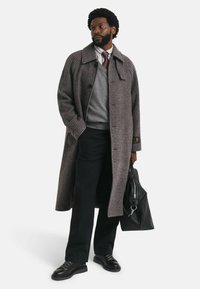 Lang, grijsbruin houndstooth-jas met een kraag, gedragen over een grijze trui en een witte gestreepte blouse; zwarte broek en schoenen; draagt een zwarte tas.
