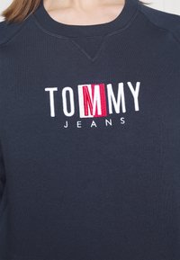 Felpa blu navy con collo rotondo a coste. Sul davanti è ricamato "TOMMY" in lettere bianche e rosse, con la scritta "JEANS" in piccolo testo nero sotto.