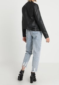 Personne portant une veste en cuir noire, un jean léger bleu déchiré avec des ourlets effilochés, et des bottines à talons noires, se tenant contre un fond blanc.