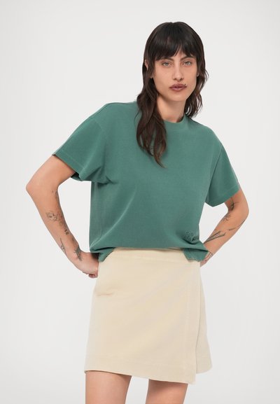 Πράσινο βαμβακερό t-shirt με κοντά μανίκια και λαιμόκοψη, συνδυασμένο με μπεζ μίνι φούστα. Διακρίνονται ορατά τατουάζ στα χέρια του μοντέλου.