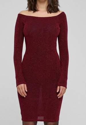 Kobieta w dopasowanej, odkrywającej ramiona, długorękawowej, bordowej sukience z brokatem na jednolitym szarym tle.