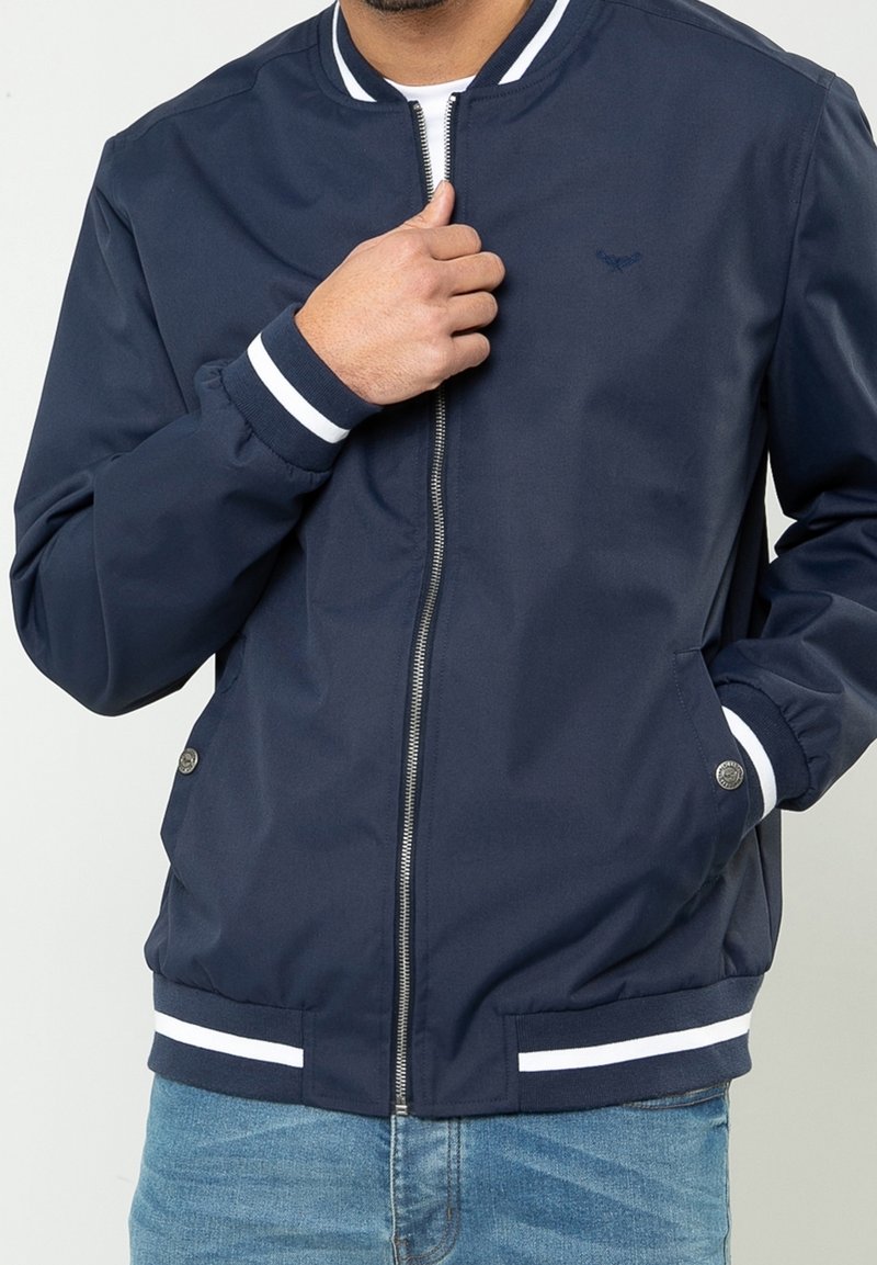 Homme portant une veste bomber bleu marine avec des poignets et une taille rayés blancs, partiellement zippée, et un jean bleu clair.