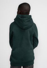 Tummanvihreä huppari, jossa on suuri kiinnitetty huppu, valmistettu pehmeästä kankaasta, rento leikkaus ja ribbikujat, nähtynä takaa.