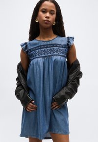 Kobieta ubrana w niebieską, haftowaną sukienkę dżinsową z falbanami na rękawach, złote kolczyki koła i czarną skórzaną kurtkę zarzuconą na ramiona.