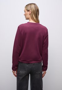 Burgunderfärgad sweatshirt med rund halsringning, ribbat avslut och långa ärmar, bärs med grå byxor. Slät tygtextur, avslappnad passform.