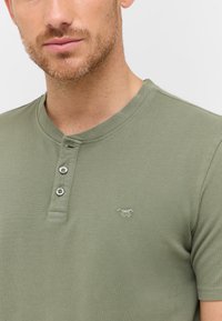 Olivenfarvet henley skjorte i et struktureret stof, med rund halsudskæring, tre knapper og et lille broderet logo på venstre bryst.