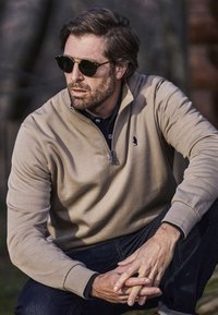 Szakállas férfi napszemüveggel, bézs negyedig cipzáras pulóvert és sötét farmert viselve, kint ül, kezeit összekulcsolva.