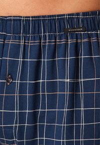 Calzoncillos tipo boxers azul marino con un patrón de cuadros, que presentan líneas blancas y marrones, una cinturilla elástica y un detalle de un solo botón. Etiqueta de la marca presente.