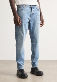 Jeans de mezclilla azul claro con corte recto, leve desgastado y diseño de cinco bolsillos, combinados con zapatos negros tipo slip-on.