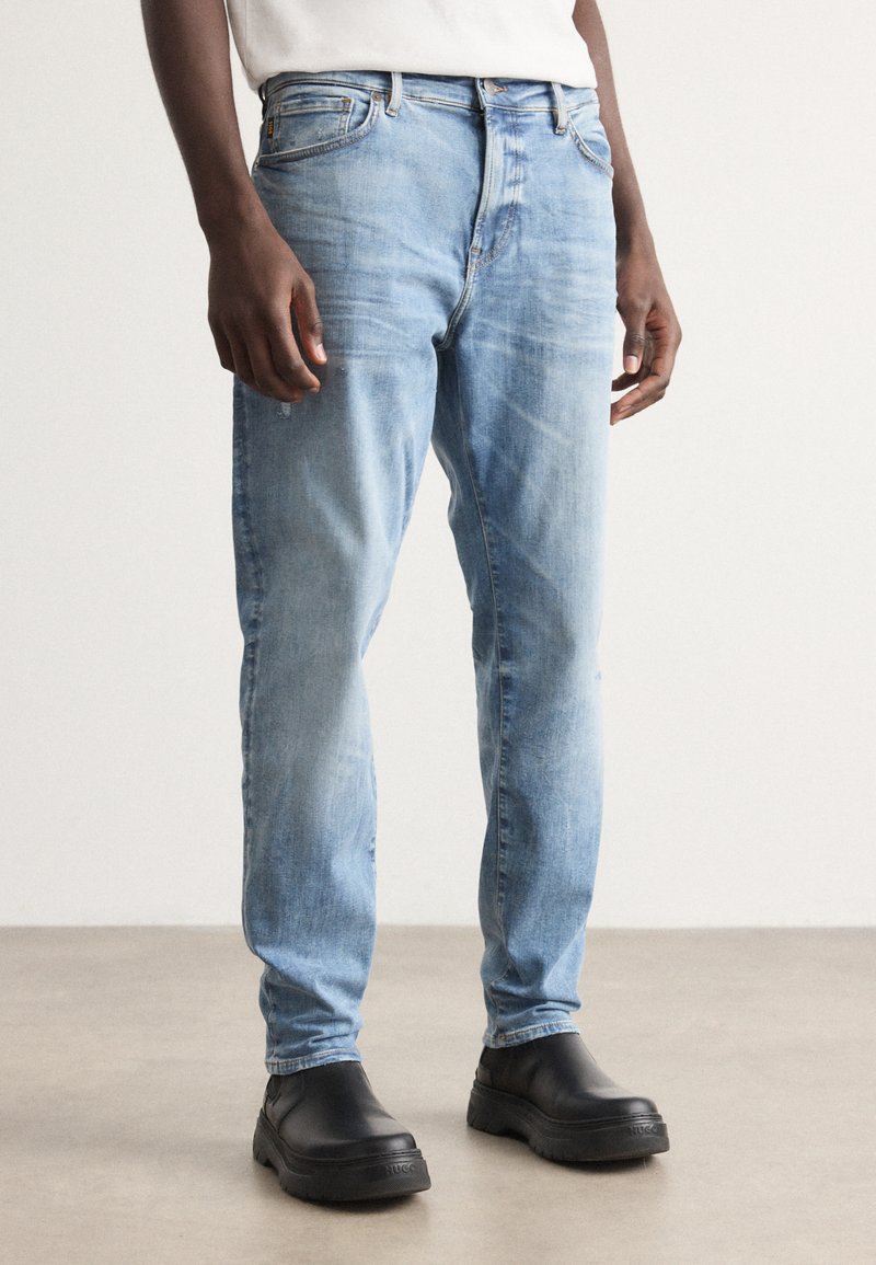 Jeans de mezclilla azul claro con corte recto, leve desgastado y diseño de cinco bolsillos, combinados con zapatos negros tipo slip-on.